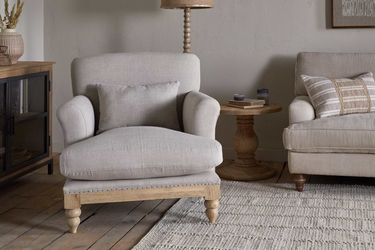 A cozy living room featuring the Kashi Linen Armchair - Natural by nkuku with a matching pillow, a round solid mango wood side table, part of a beige sofa, a textured rug, and wood flooring in neutral decor.