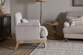 A cozy living room featuring the nkuku Kashi Linen Armchair in natural beige, a matching sofa with striped cushions, a round solid mango wood side table, floor lamp, and cabinet on a light woven rug over wooden floors.