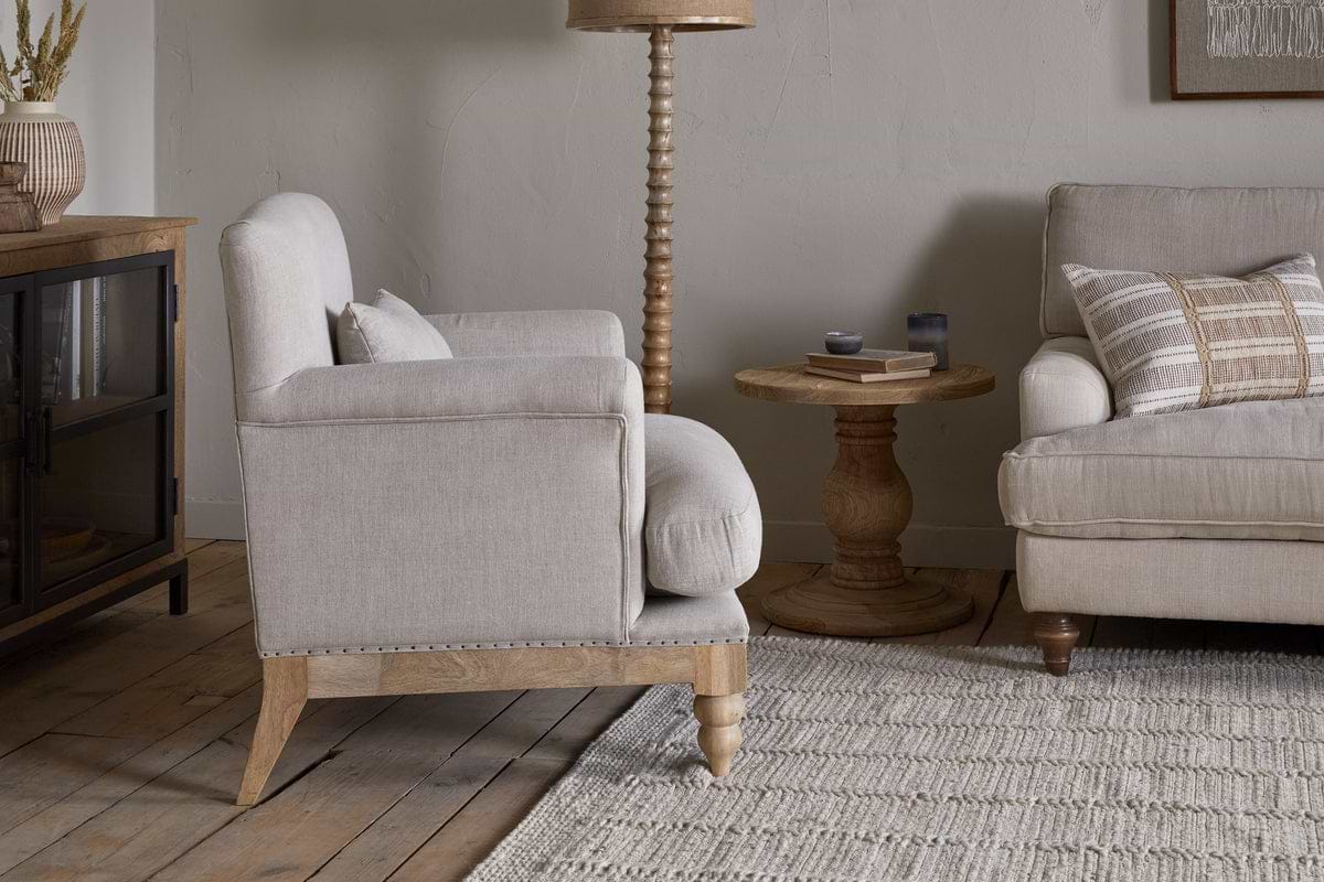 A cozy living room featuring the nkuku Kashi Linen Armchair in natural beige, a matching sofa with striped cushions, a round solid mango wood side table, floor lamp, and cabinet on a light woven rug over wooden floors.
