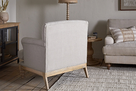 A neutral-toned living room features the nkuku Kashi Linen Armchair in natural beige, a matching sofa with striped cushions, a solid mango wood side table, woven rug, and decor on a cabinet set against a light wall.
