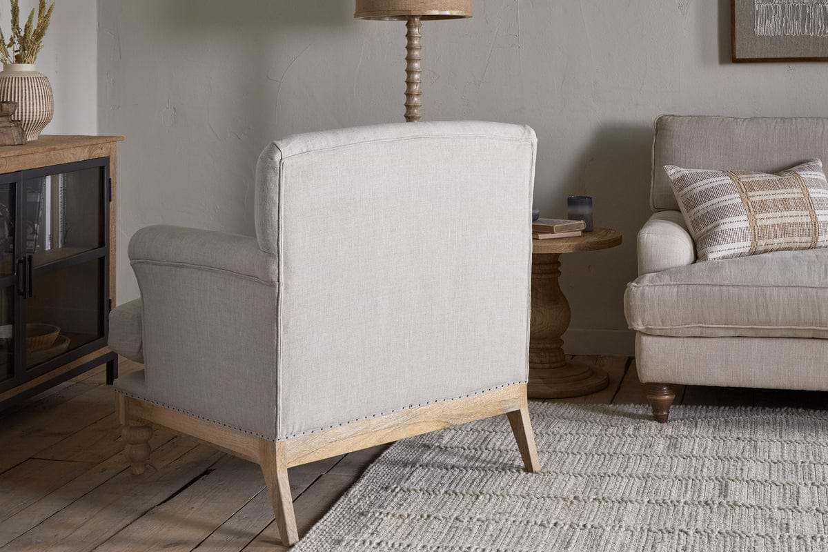 A neutral-toned living room features the nkuku Kashi Linen Armchair in natural beige, a matching sofa with striped cushions, a solid mango wood side table, woven rug, and decor on a cabinet set against a light wall.