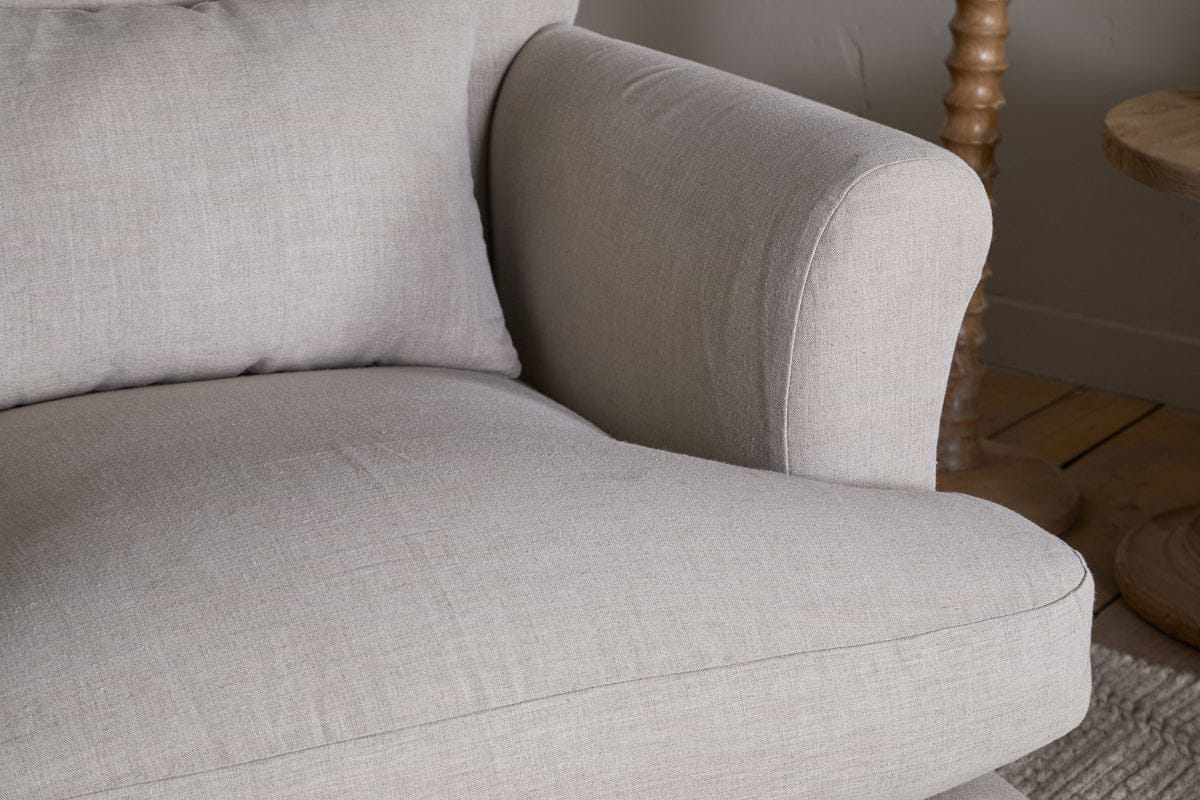 Close-up of the arm and cushion of the Kashi Linen Armchair - Natural by nkuku, upholstered in light beige linen, with a round solid mango wood side table and wooden floor visible in the background.
