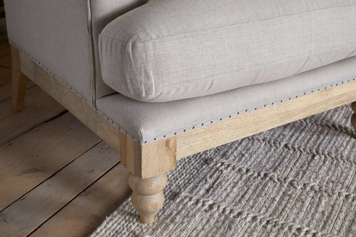 Close-up of the nkuku Kashi Linen Armchair in Natural, featuring beige upholstery and solid mango wood frame and legs, set on a textured, light-colored woven rug atop a wooden floor.