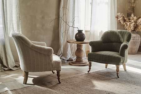 Two nkuku Kiana Cotton Armchairs in natural face a hand-carved mango wood side table topped with a dark vase and bare branch. Sunlight through sheer curtains highlights the room’s neutral, rustic, and cozy feel with natural textures.