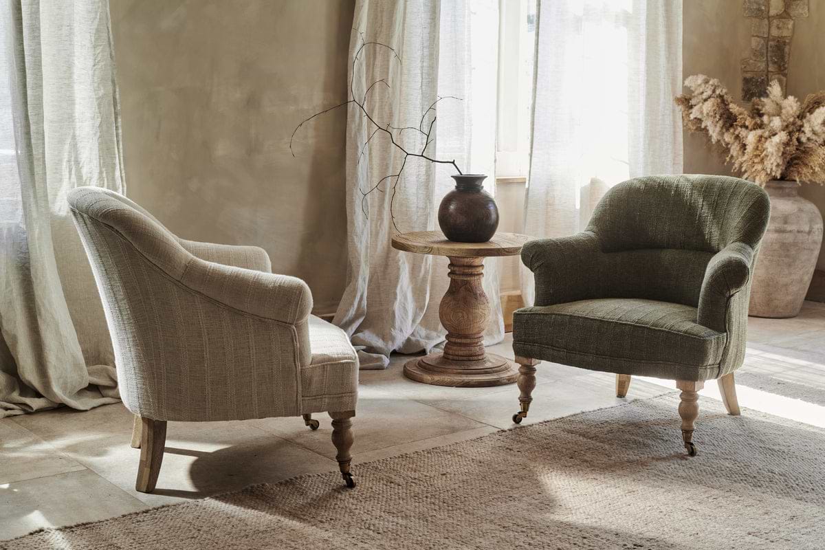 Two nkuku Kiana Cotton Armchairs in natural face a hand-carved mango wood side table topped with a dark vase and bare branch. Sunlight through sheer curtains highlights the room’s neutral, rustic, and cozy feel with natural textures.