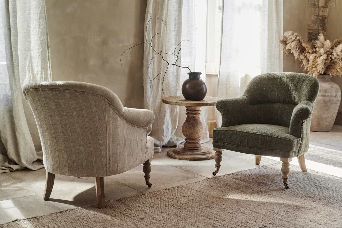 Two Kiana Cotton Armchairs by nkuku are arranged around a hand-carved mango wood table with a dark vase in a softly lit, neutral room featuring sheer curtains and rustic decor with dried grasses in the background.