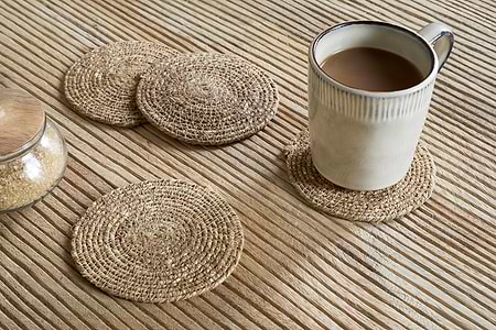 A coffee-filled mug rests on an nkuku Keso Woven Coaster - Brown (Set of 4), with three more coasters and a glass jar of brown sugar with a wooden lid arranged on the textured wooden surface.