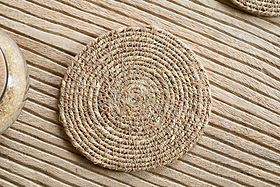 A Keso Woven Coaster - Brown from nkuku rests on a light wooden, striped surface, with part of a grain-filled glass container on the side, highlighting the minimalist style of this handwoven tableware set.