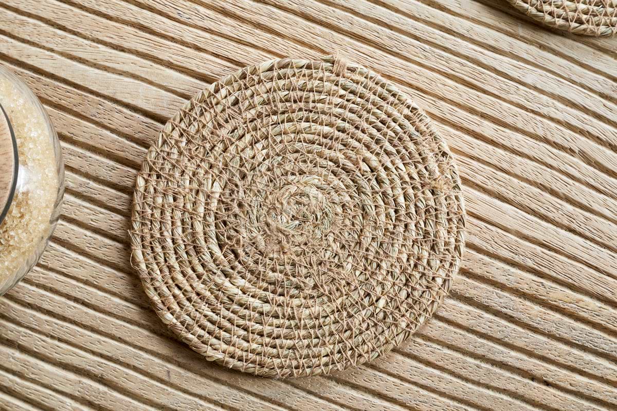 A Keso Woven Coaster - Brown from nkuku rests on a light wooden, striped surface, with part of a grain-filled glass container on the side, highlighting the minimalist style of this handwoven tableware set.