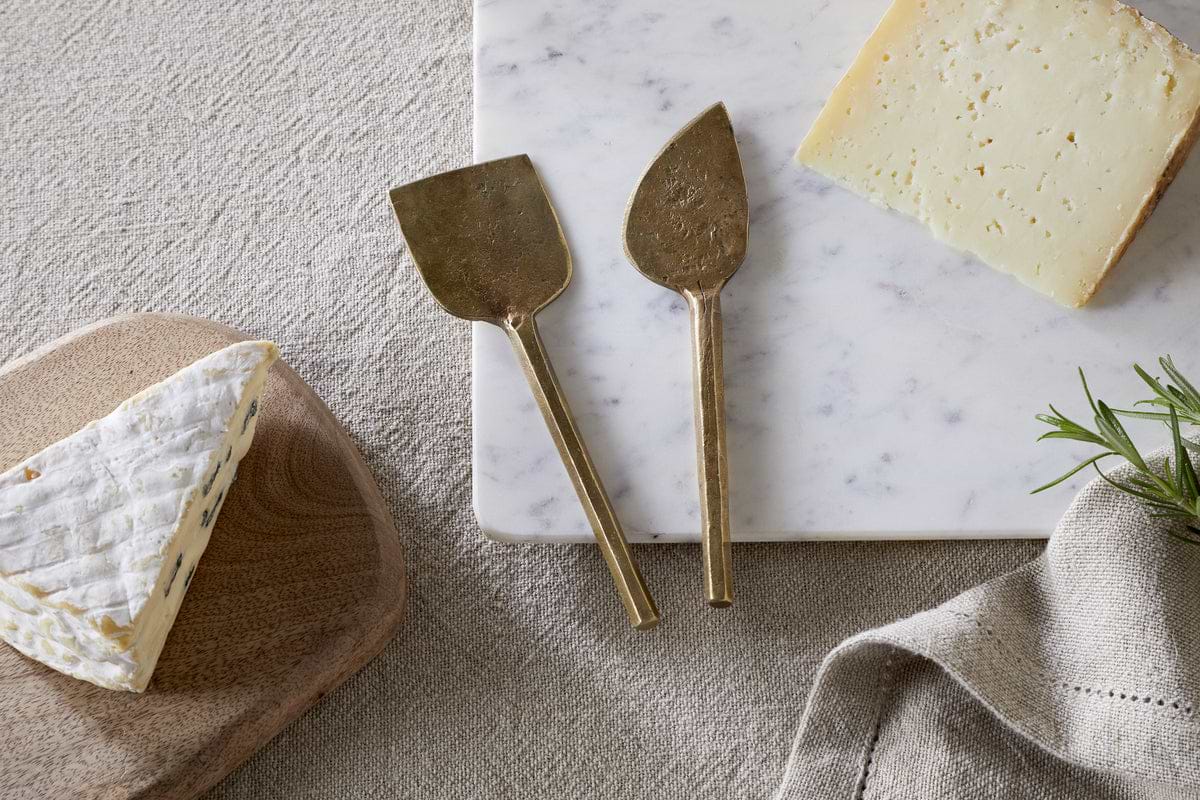 The nkuku Kasa Cheese Knives - Antique Brass (Set of 2) are displayed on a white marble board with hard cheese and brie on a wooden board, accompanied by a beige napkin and rosemary.