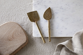 The nkuku Kasa Cheese Knives—antique brass, set of 2—rest on a white marble cheese board beside a wooden board and beige linen cloth, all arranged on a textured, light surface.