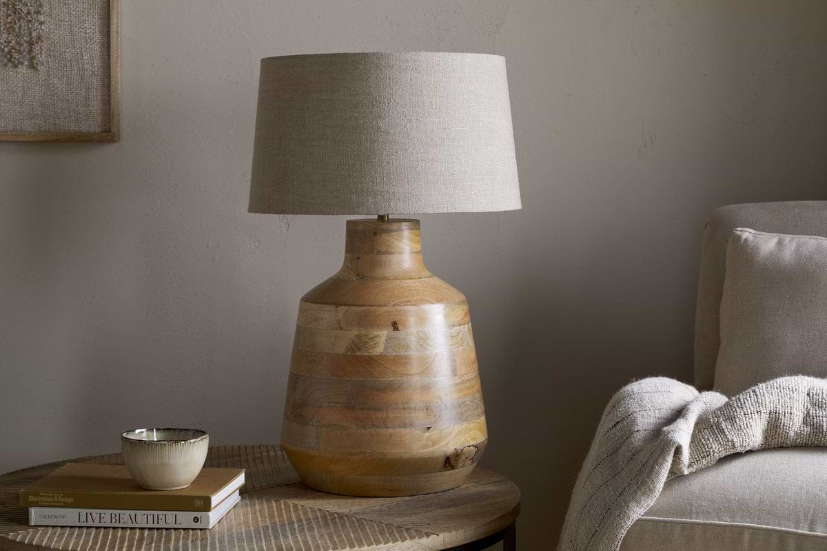 The nkuku Kayo Statement Table Lamp Mango Wood - Natural, handcrafted in India with a wooden base and beige shade, sits on a round side table beside books and décor—perfect statement lighting for cozy corners.