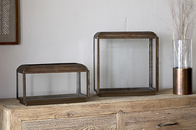 Two glass and metal display cases—one rectangular, one square—sit on a wooden cabinet. The scene is cozily lit by nkuku’s Kiwanda Tall Lantern in antique brass, with dried stems and artwork set against a soft light-colored wall.