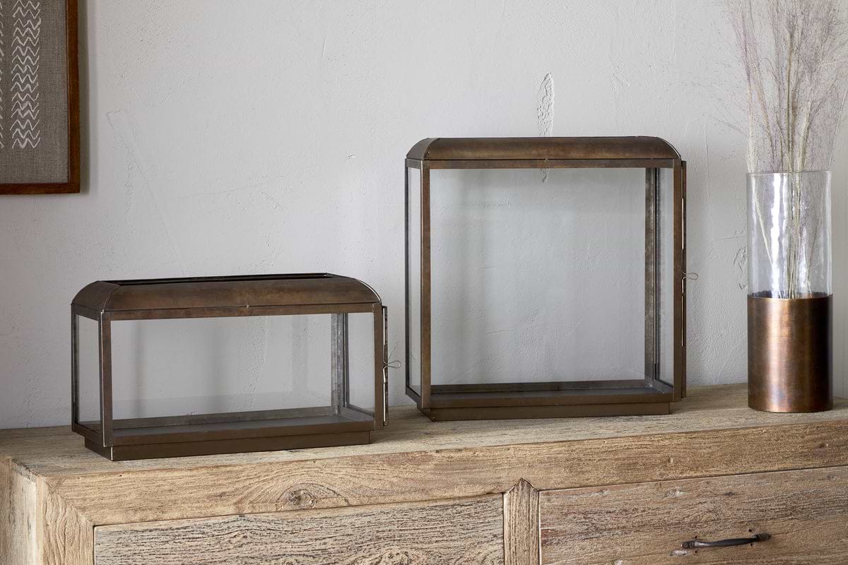 Two glass and metal display cases—one rectangular, one square—sit on a wooden cabinet. The scene is cozily lit by nkuku’s Kiwanda Tall Lantern in antique brass, with dried stems and artwork set against a soft light-colored wall.