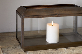 The Kiwanda Tall Lantern - Antique Brass by nkuku features a white pillar candle burning inside its rectangular metal and glass frame, adding timeless charm to any wooden surface and creating elegant candlelight against light-colored walls.