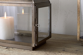 A white pillar candle glows inside the nkuku Kiwanda Tall Lantern in Antique Brass, adding candlelit decor to a wooden surface against a light-colored wall.