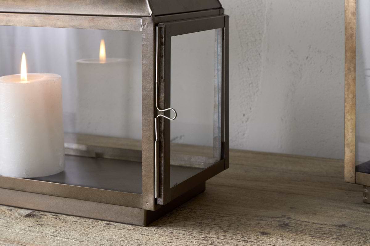 A white pillar candle glows inside the nkuku Kiwanda Tall Lantern in Antique Brass, adding candlelit decor to a wooden surface against a light-colored wall.