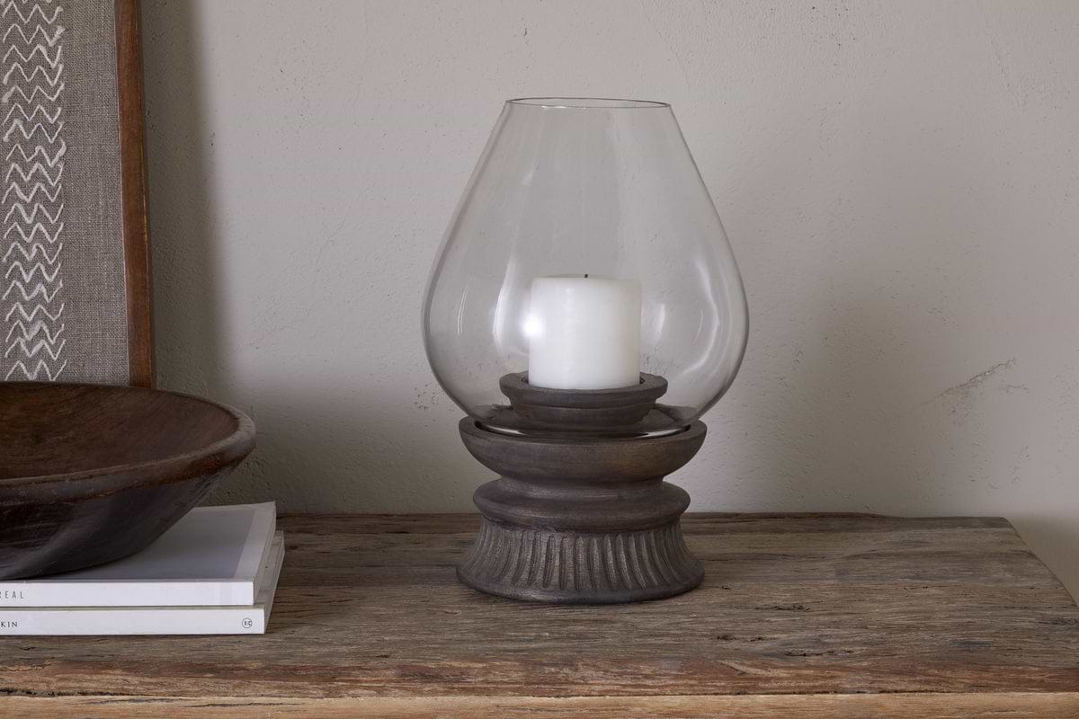 A white rustic pillar candle is placed inside the nkuku Kawani Mango Wood Lantern - Dark Ash on a wooden table, accompanied by stacked books and a wooden bowl.