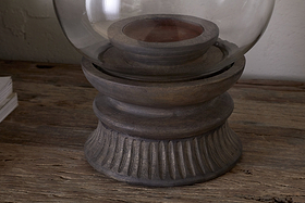 A close-up of the nkuku Kawani Mango Wood Lantern in Dark Ash, featuring rustic carved mango wood and a glass dome, sits on a weathered wooden surface beside a small stack of white paper.
