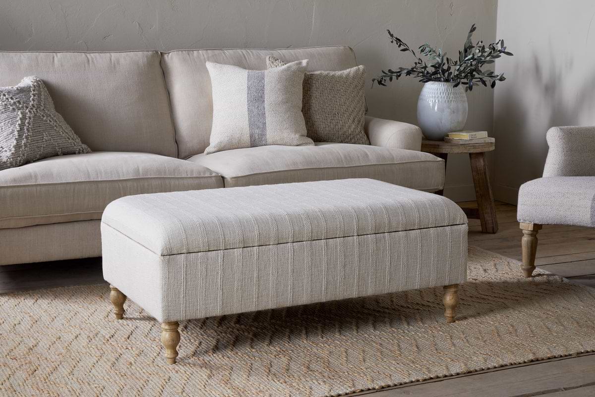 The Kiana Upholstered Storage Ottoman by nkuku in natural sits on a woven rug before a light sofa with pillows. Nearby, a side table holds a vase of greenery, completing the neutral-toned living space.