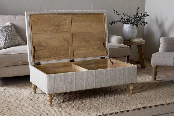 The Kiana Upholstered Storage Ottoman - Natural by nkuku, with wooden legs and two storage compartments, sits open on a woven rug in the living area. In the background are a couch, small table, vase with greenery, and hardcover book.