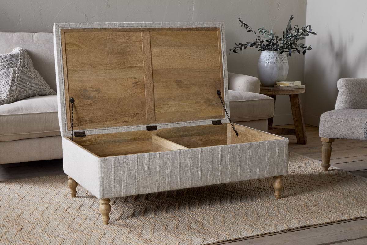 The Kiana Upholstered Storage Ottoman - Natural by nkuku, with wooden legs and two storage compartments, sits open on a woven rug in the living area. In the background are a couch, small table, vase with greenery, and hardcover book.