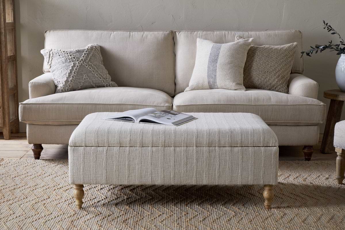 A beige sofa with four textured throw pillows sits behind the Kiana Upholstered Storage Ottoman by nkuku in Natural, which holds an open book on top, all arranged on a woven rug in a cozy, neutral-toned living space.