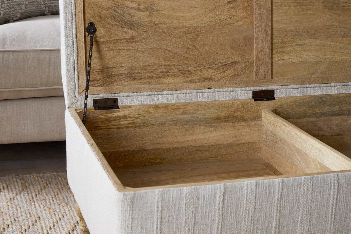 A close-up of the nkuku Kiana Upholstered Storage Ottoman - Natural shows its cushioned fabric exterior, wooden interior with hinge mechanism and divider, set beside a beige sofa and textured rug in a living space.