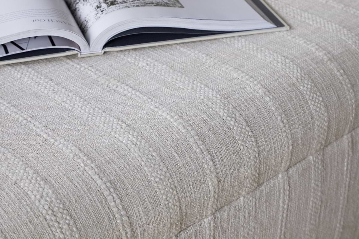 A close-up of the nkuku Kiana Upholstered Storage Ottoman in Natural, featuring a cream-striped, textured fabric and cushioned top, with an open magazine resting on it—an inviting accent for any living space.
