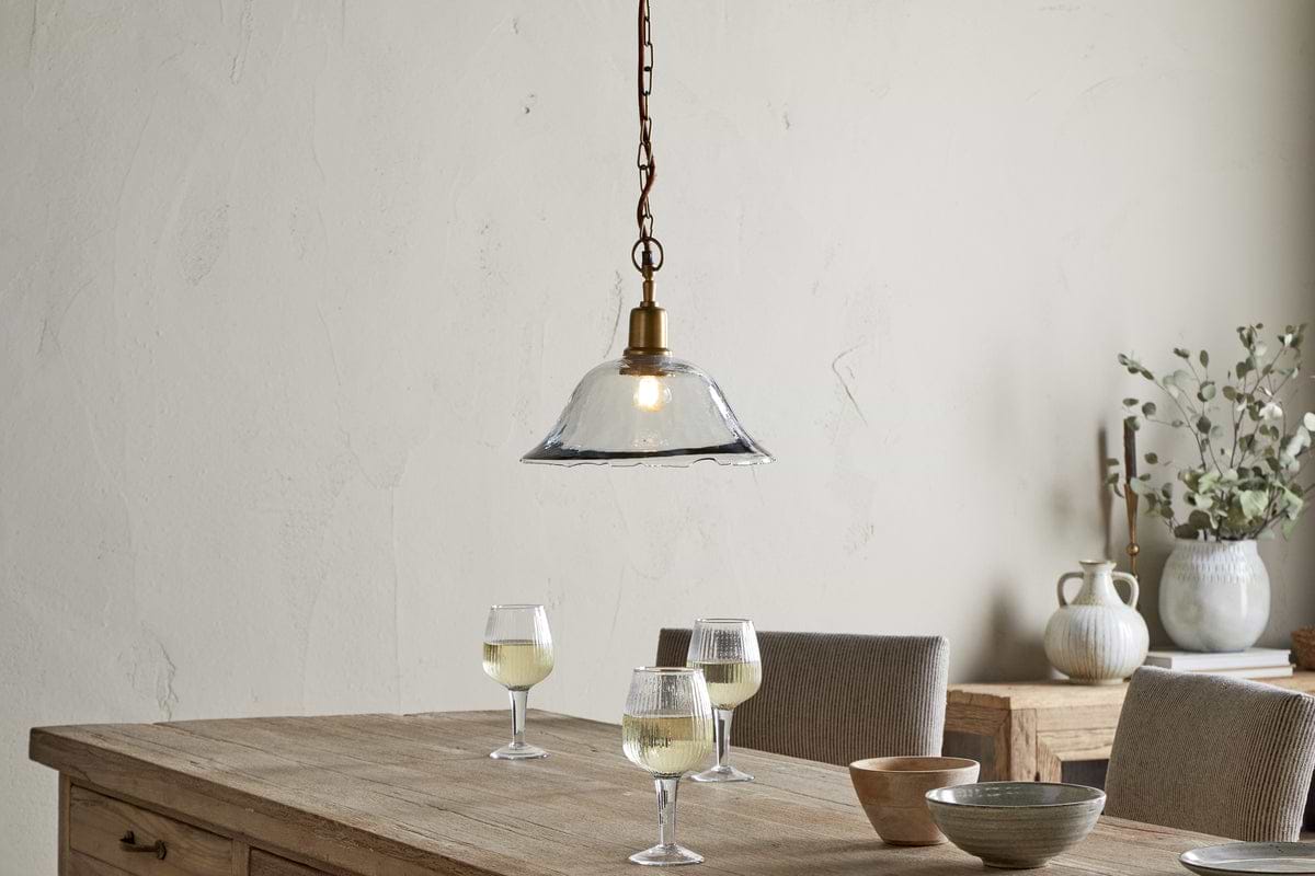 A rustic wooden table with three white wine glasses, ceramic and wooden bowls sits under the nkuku Keju Organic Shape Recycled Glass Pendant - Clear. Behind are cushioned chairs and a sideboard with vases and greenery against a neutral wall.