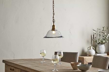 A rustic wooden table with three white wine glasses, ceramic and wooden bowls sits under the nkuku Keju Organic Shape Recycled Glass Pendant - Clear. Behind are cushioned chairs and a sideboard with vases and greenery against a neutral wall.
