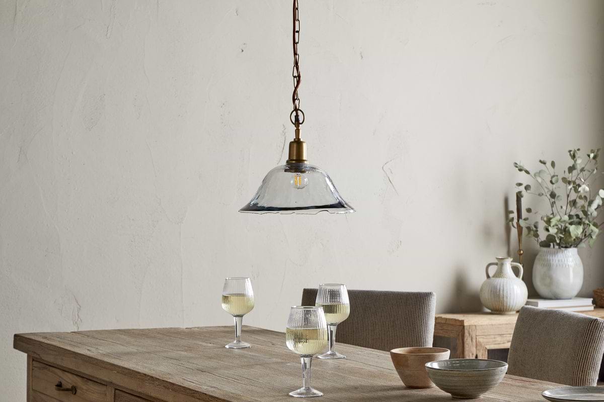 A rustic wooden table holds three glasses of white wine, a small bowl, and a wooden dish. Above, the nkuku Keju Organic Shape Recycled Glass Pendant in clear hangs from a chain. Neutral pottery and greenery decorate the sideboard against a beige wall.