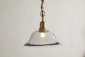 The Keju Organic Shape Recycled Glass Pendant - Clear by nkuku features a wavy-edged glass shade and antique brass fixture, suspended from a twisted cord. Its softly glowing bulb casts gentle reflections, enhancing minimalist, vintage decor.