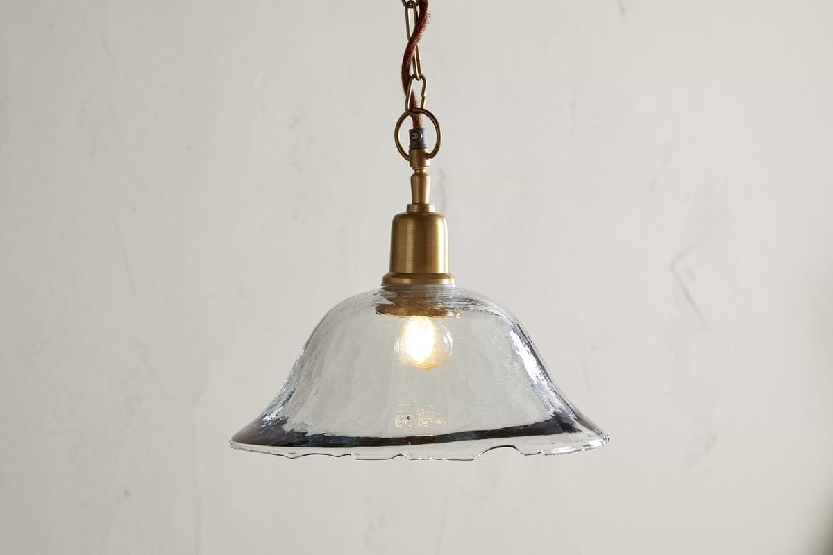 The Keju Organic Shape Recycled Glass Pendant - Clear by nkuku features a wavy-edged glass shade and antique brass fixture, suspended from a twisted cord. Its softly glowing bulb casts gentle reflections, enhancing minimalist, vintage decor.