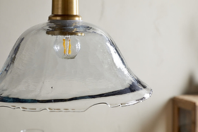 A close-up of the nkuku Keju Organic Shape Recycled Glass Pendant in clear, featuring a wavy glass shade, visible filament bulb, and antique brass stem, set against a light-textured wall with part of a wooden object.
