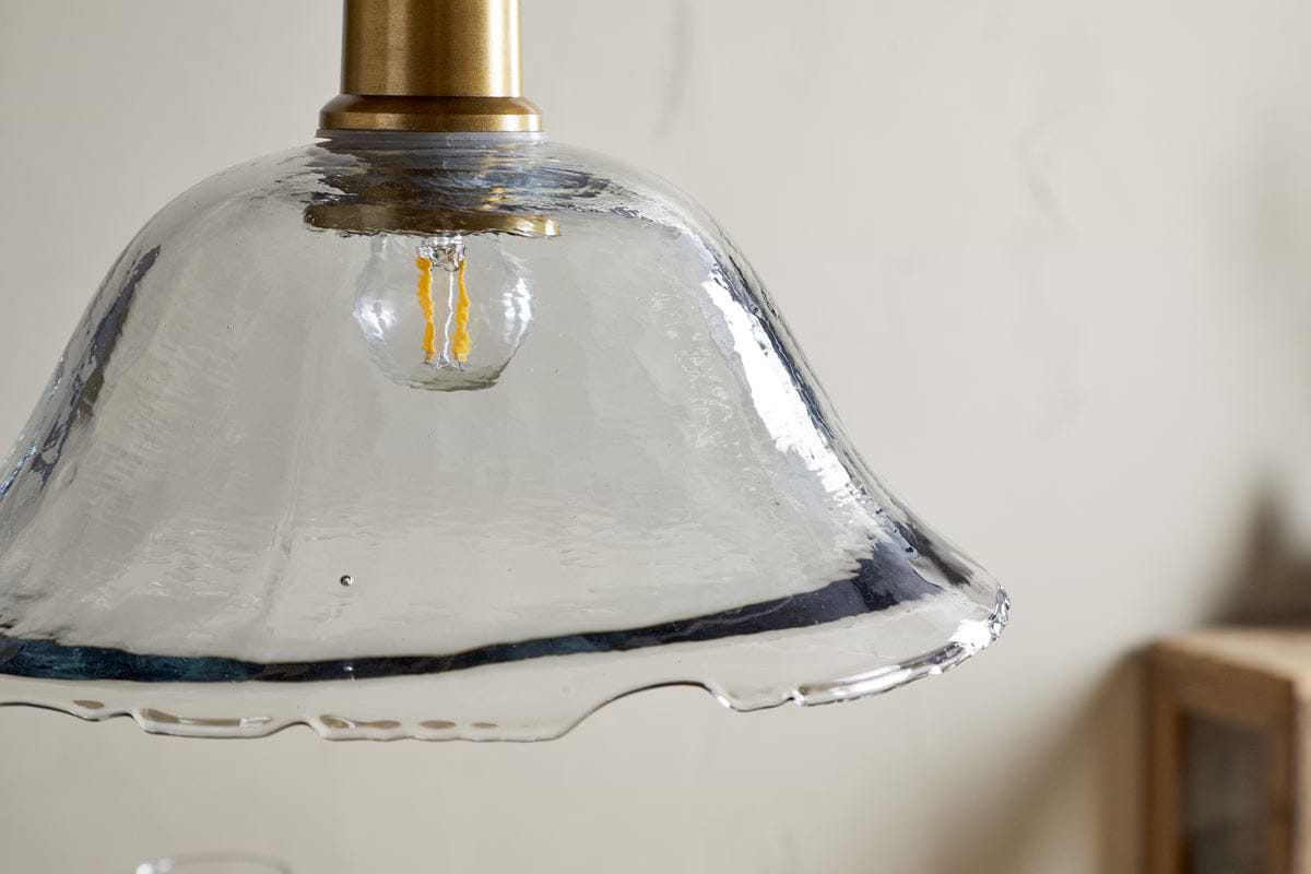 A close-up of the nkuku Keju Organic Shape Recycled Glass Pendant in clear, featuring a wavy glass shade, visible filament bulb, and antique brass stem, set against a light-textured wall with part of a wooden object.