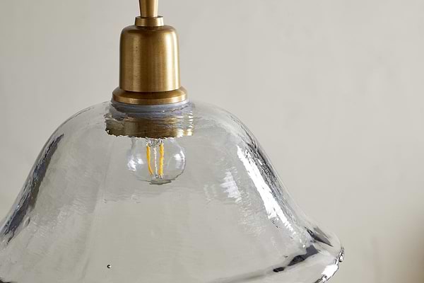 A close-up of the Keju Organic Shape Recycled Glass Pendant - Clear by nkuku shows its wavy, transparent glass shade and antique brass top, with a small LED filament bulb inside, emphasizing the lamp’s unique texture and eco-friendly materials.