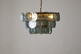 The Kagitha Recycled Glass Chandelier - Green by nkuku features three tiers of overlapping, round, translucent green-tinted glass discs hanging from a gold chain, showcasing a striking layered design against an off-white wall.