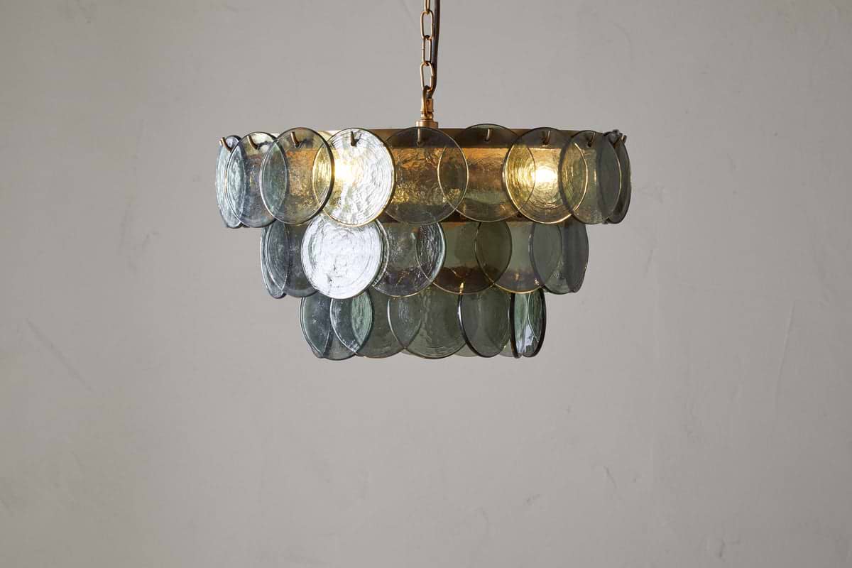 The Kagitha Recycled Glass Chandelier - Green by nkuku features three tiers of overlapping, round, translucent green-tinted glass discs hanging from a gold chain, showcasing a striking layered design against an off-white wall.