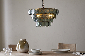 A modern dining table with two wooden chairs, set with a beige vase, stacked ceramic bowls and plates, two clear glasses, and a cloth napkin. Above hangs the nkuku Kagitha Recycled Glass Chandelier in green, softly illuminating the scene.