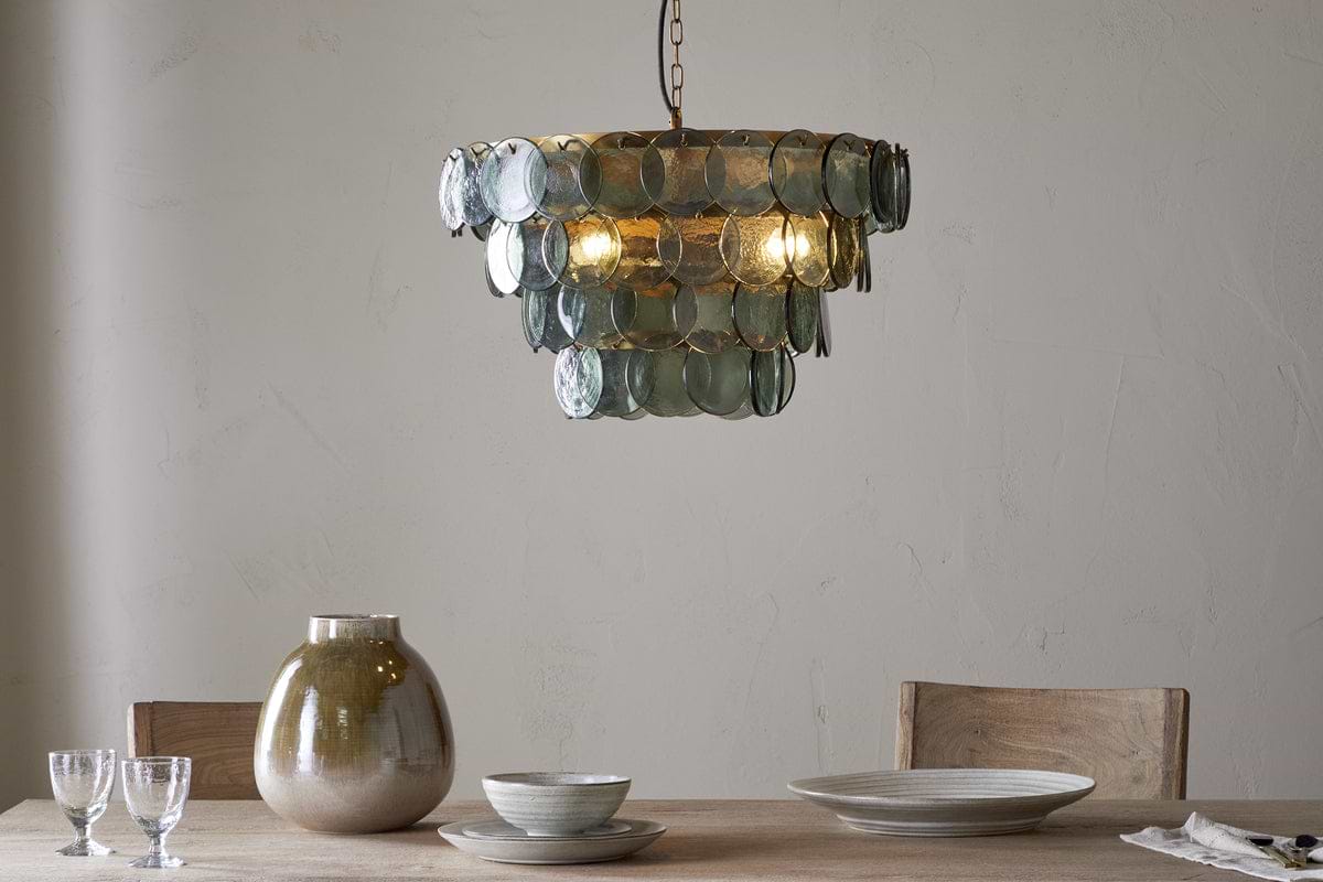 A modern dining table with two wooden chairs, set with a beige vase, stacked ceramic bowls and plates, two clear glasses, and a cloth napkin. Above hangs the nkuku Kagitha Recycled Glass Chandelier in green, softly illuminating the scene.