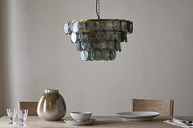 A rustic dining table with light wood chairs holds ceramic dishes, two clear glasses, and a large beige vase. Above hangs the nkuku Kagitha Recycled Glass Chandelier - Green, its layered design casting a soft, elegant glow.