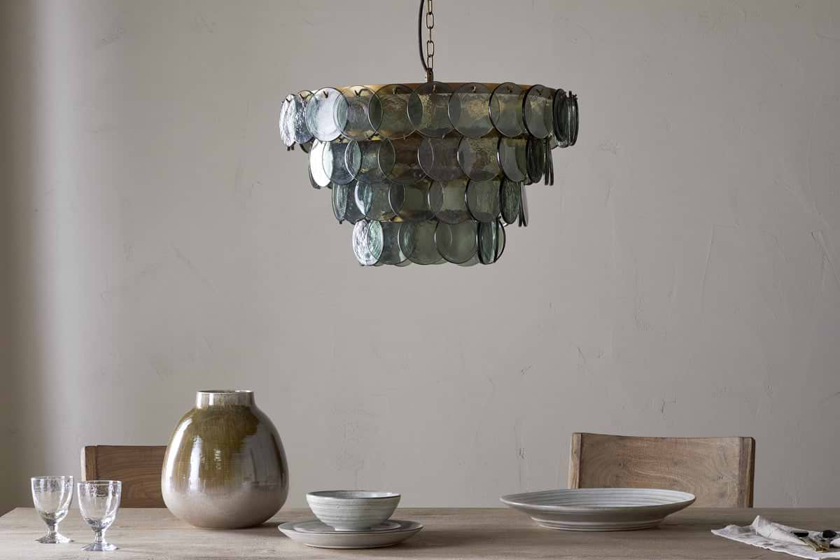 A rustic dining table with light wood chairs holds ceramic dishes, two clear glasses, and a large beige vase. Above hangs the nkuku Kagitha Recycled Glass Chandelier - Green, its layered design casting a soft, elegant glow.