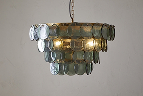 The Kagitha Recycled Glass Chandelier - Green by nkuku features two tiers of overlapping, round, translucent green glass discs and elegant brass detailing, casting a soft glow and subtle reflections on a plain white wall.