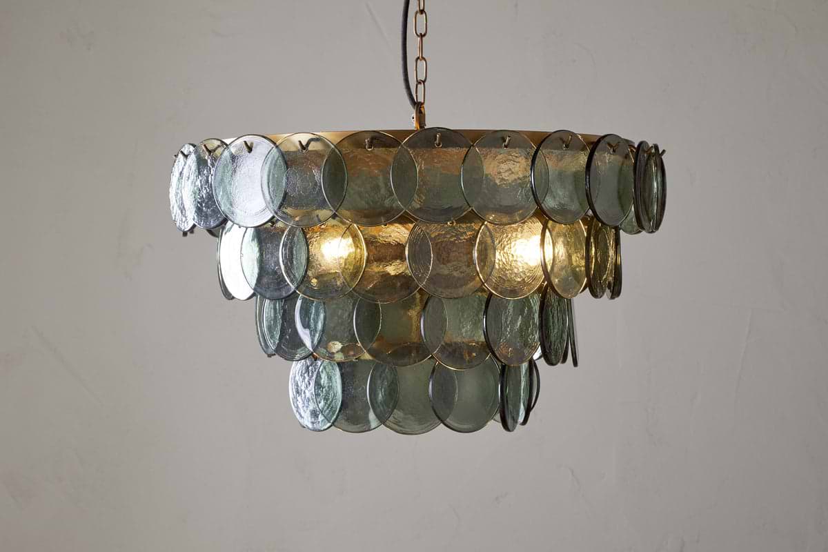 The Kagitha Recycled Glass Chandelier - Green by nkuku features two tiers of overlapping, round, translucent green glass discs and elegant brass detailing, casting a soft glow and subtle reflections on a plain white wall.