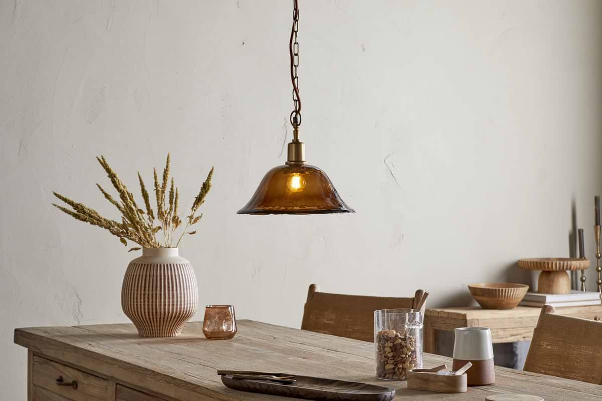 A rustic dining table sits beneath the nkuku Keju Organic Shape Recycled Glass Pendant in Dark Amber, its warm glow highlighting a ribbed vase of dried grasses, small bowls, glassware, and a wooden tray in a softly lit, minimalist room.