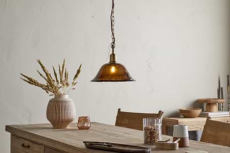 A rustic dining table sits beneath the nkuku Keju Organic Shape Recycled Glass Pendant in Dark Amber, its warm glow highlighting a ribbed vase of dried grasses, small bowls, glassware, and a wooden tray in a softly lit, minimalist room.