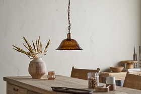 A rustic dining table with a ribbed vase of dried grasses, the nkuku Keju Organic Shape Recycled Glass Pendant in Dark Amber above, and assorted dishes and decor, all set against a neutral textured wall.