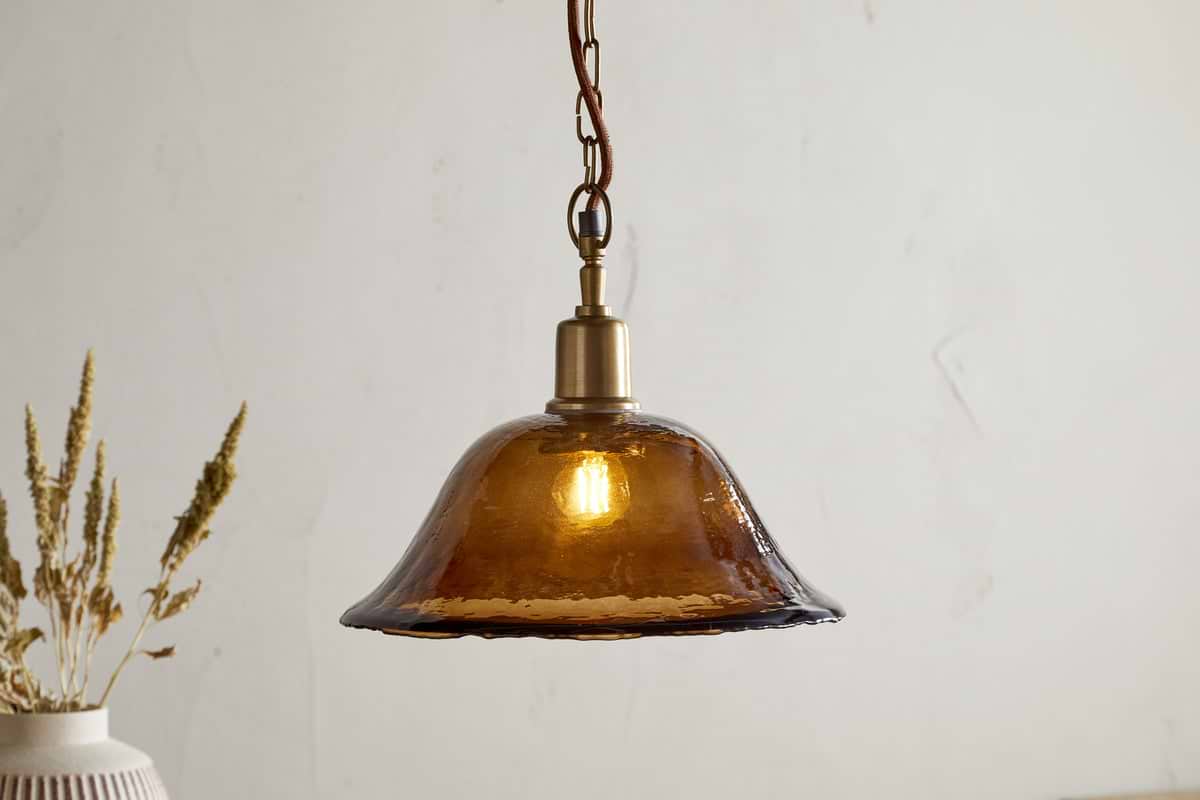 The Keju Organic Shape Recycled Glass Pendant in dark amber by nkuku features a visible bulb and antique brass fixture suspended from a chain. In the background, dried flowers in a vase rest against a light-colored wall.