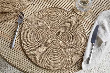 A Keso Woven Placemat by nkuku sits on a light wooden table, paired with minimalist tableware and a glass of water on a handwoven coaster in the upper right corner.